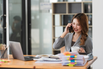 Confident Asian businesswoman using smartphone and drinking coffee at office desk with laptop and documents.