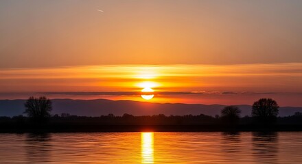 Obraz premium Serene sunset over calm lake waters with trees silhouetted against vibrant orange sky
