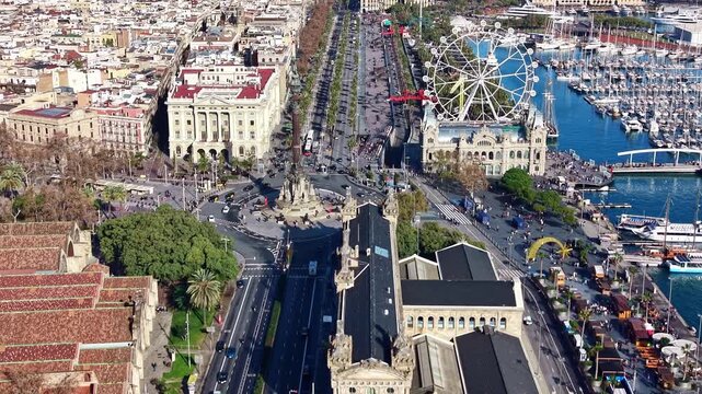 High aerial drone shot overlooking the Columbus Monument at the end of La Rambla, with views of the Ferris wheel, Rambla de Mar, and the entire Port Vell marina