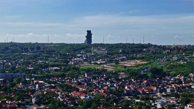 Sweeping aerial panorama reveals iconic Garuda Wisnu Kencana GWK statue towering above Kuta Selatan residential rooftops green hills and telecom towers under clear blue sky Bali