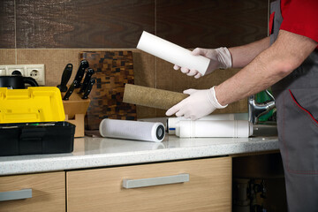 A plumber holds a used water filter cartridge and new cartridge in front of a home kitchen.