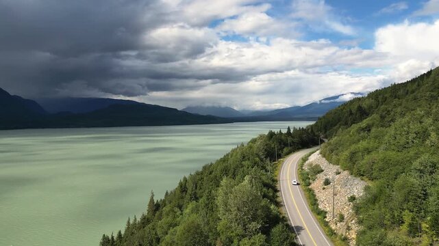 Aerial drone flies along the shoreline with the white van farther in the distance, a more prominent lake surface illuminated by sunlight, and rainclouds gathering over distant mountains in British Col