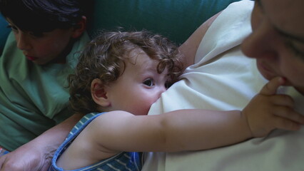 Breastfeeding toddler looks up at mother while nursing on couch, close sibling nearby, intimate family moment of bonding and nourishment indoors