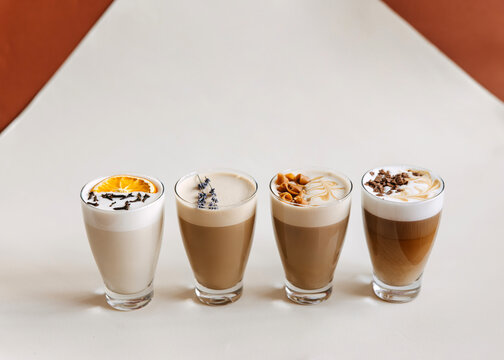 Four artisanal latte and coffee drinks in clear glasses with decorative foam toppings, arranged in a minimalist studio setting. Modern beverage presentation with warm tones and clean background.