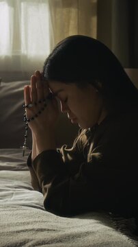 Vertical shot of gen Z girl holding rosary with cross finishing her morning prayer and lowering head while connecting with god