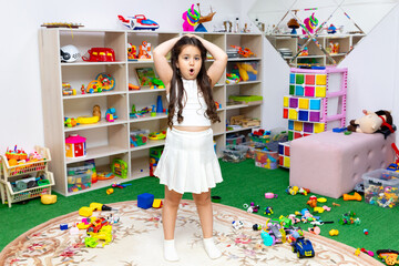 Surprised little girl in white outfit stands in messy children room. Kid playing among the many...