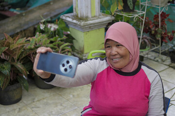 Naklejka premium Middle-aged Asian woman wearing a hijab takes a selfie on her home terrace after completing a workout. Fitness, healthy lifestyle, and home exercise concept.