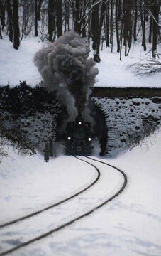 View of a vintage steam train emerges from a dark tunnel amidst a snowy forest landscape, the black smoke contrasting with the white snow, Budapest, Hungary.