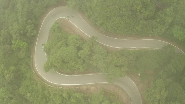 Aerial view of a black car navigating a sharp bend on a winding road cutting through lush green trees in Sakleshpura, Sakleshpura, Karnataka, India.