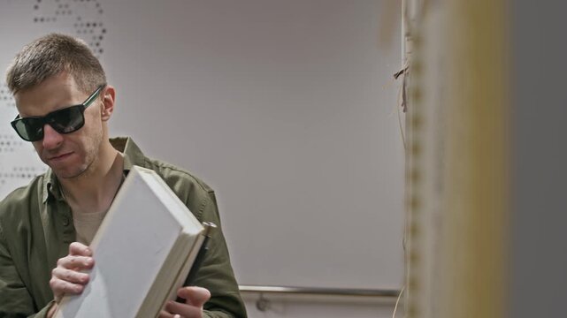 Chest up shot of focused middle-aged Caucasian man with visual impairment choosing book from shelf in modern well-equipped library for blind people, reading braille covers with fingers