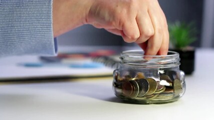 Hand dropping coins into glass savings jar on table. Close up of fingers inserting money into transparent container, representing simple financial planning and goal achievement in everyday life