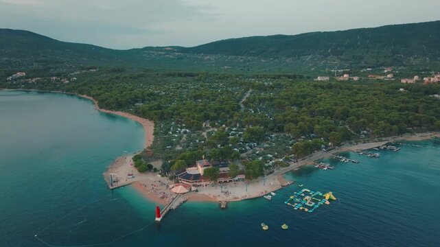Aerial view of a vibrant coastline with a striking red lighthouse contrasting against the turquoise sea and lush green forests, Cres, Primorsko-goranska zupanija, Croatia.