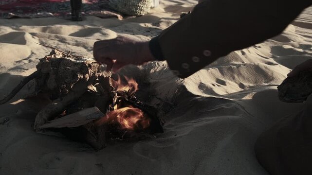 Hand of man dressed in traditional berber clothes tending small open fire in desert sand. Nomadic lifestyle, cultural ritual, survival practice in arid environment. Sahara desert, Siwa oasis, Egypt