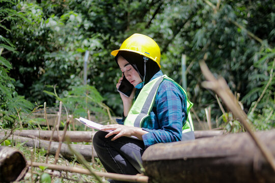 Asian woman wearing a yellow hard hat and safety vest talks on her phone while writing notes on a clipboard in a forest. Field inspection, environmental work, and professional documentation concept