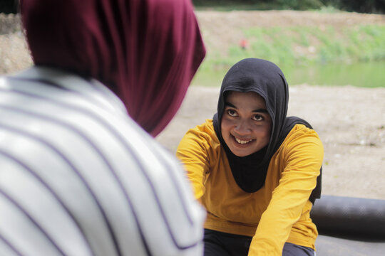 Two young Asian women wearing hijabs practice yoga outdoors, with one woman assisting the other in a stretching pose. Mindfulness, Wellness, flexibility, support, and healthy lifestyle concept