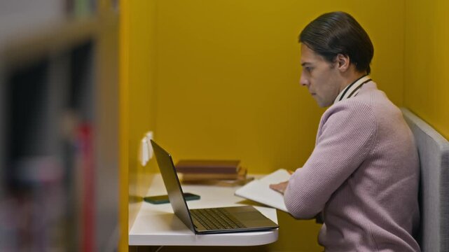 Side view shot of concentrated young Caucasian male student sitting in front of laptop and making notes while working on thesis or dissertation in modern library