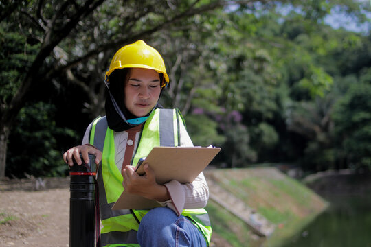 Young Asian woman engineer wearing a hijab, hard hat, and safety vest reviews project documents on a clipboard while holding a blueprint tube outdoors. Civil engineering, project planning concept.
