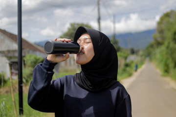 Young Asian woman wearing a hijab drinks water from a bottle while taking a break during an outdoor run. Hydration, fitness, workout routine, and healthy active lifestyle concept