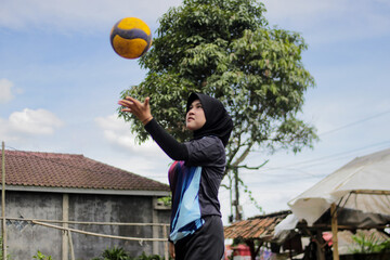 Young Asian woman volleyball player wearing a hijab practices serving on an outdoor volleyball court in a rural village setting. Sports training, grassroots athletics, and active lifestyle concept