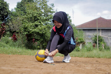 Young Asian woman volleyball player wearing a hijab ties her shoelace beside a volleyball while preparing for practice on an outdoor volleyball court in a rural village setting