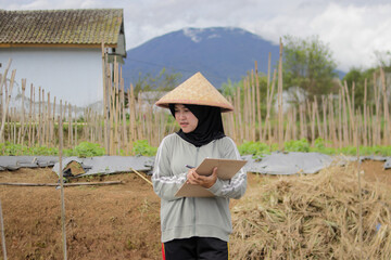 Young Asian woman farmer wearing a hijab and woven bamboo hat writes on a clipboard while observing crop growth in agricultural fields. Agriculture, rural livelihood, and outdoor lifestyle concept