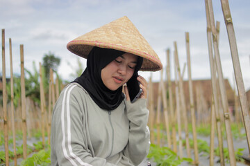 Young Asian female farmer wearing a hijab and woven bamboo hat talks on her smartphone while inspecting crop growth in agricultural fields. Modern farming, rural work, agriculture management concept