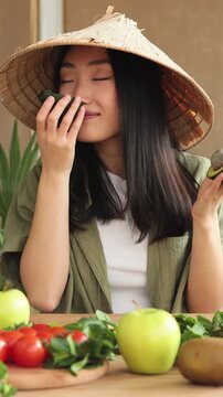 A young Asian woman wearing a conical hat holds avocados and smiles at the camera with a variety of fresh fruits and vegetables on the table