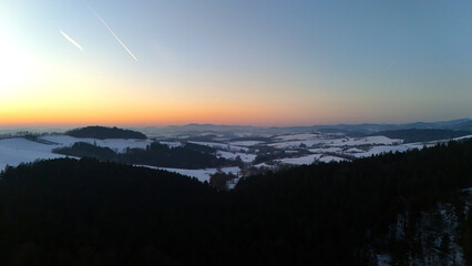 Fototapeta premium Aerial view of a tranquil winter landscape at sunset. Dark silhouettes of pine forests contrast with snow-covered rolling hills and valleys under a colorful twilight sky with airplane contrails.
