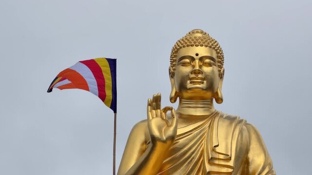 Golden seated Buddha statue displays Vitarka mudra gesture as a Buddhist flag moves beside it, with a gray overcast sky creating a contemplative visual setting. Temple icon.