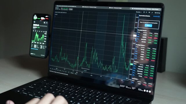 close up smartphone with laptop and open stock exchange, trading Online app interface. Man hand scrolling touch screen analysing chart forex crypto currency.