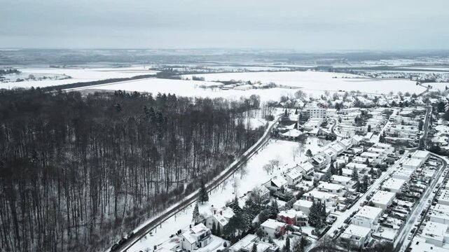 Forward aerial glide over snowy Elchingen, showcasing dense winter forest, winding roads, residential homes, and distant snowy fields under overcast sky.