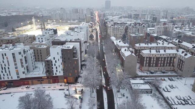 Cinematic aerial view of snow-covered urban avenue with traffic, modern buildings, and street lights during winter dusk in Paris, France