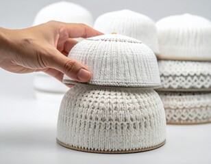 Hand holding traditional white kufi cap stacked on top of another kufi cap with soft background lighting studio shot