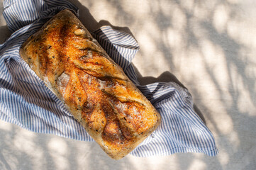 homemade multigrain sourdough bread is placed on blue stripes linen napkin with cream linen cloth covered table, in natural sunlight shining through leaves, making abstract shadows.