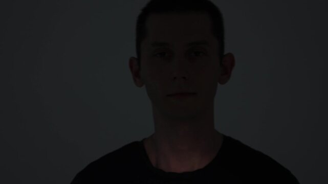 A close-up video portrait of a melancholic young man staring piercingly into the camera. Dynamic spotlight illuminates his face before fading into shadow.
