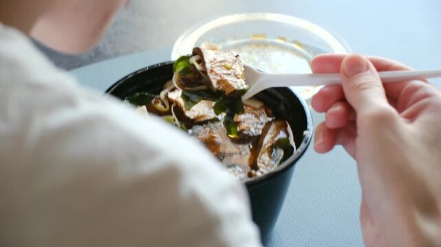 Close up view from over shoulder of woman eating hot udon noodle soup with mushrooms and seaweed from black plastic bowl, using disposable spoon for quick lunch break. Woman eat soup ramen.