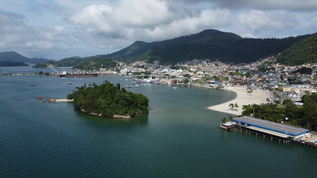 Wide coastal view of Angra dos Reis in Brazil showing sandy beach areas, urban waterfront, and tropical mountains rising behind the town. Natural scenery and city life along the Atlantic coast.
