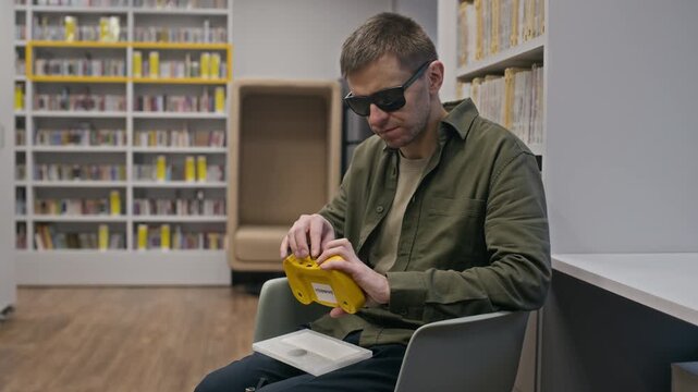 Medium long shot of middle-aged Caucasian blind man sitting on chair in modern accessible library among bookshelves and using digital audiobook player for visually impaired people