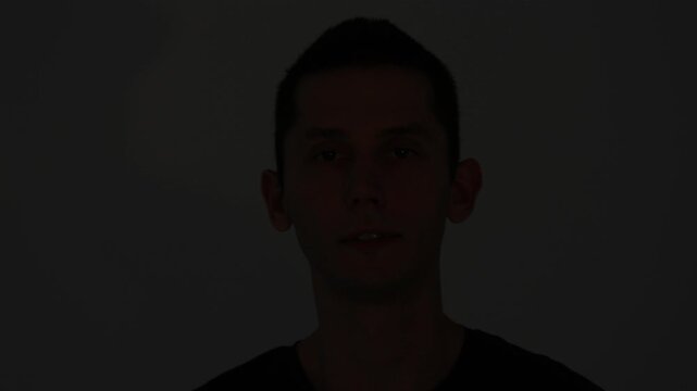 A large-scale video portrait. A young man with a piercing, melancholic look is caught in a dynamic beam of light that eventually fades, leaving him in silhouette.