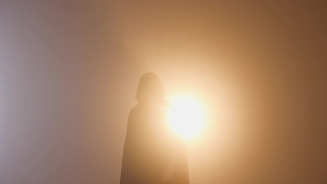 A figure in a black hooded cloak, his features hidden, performs mystical hand signs while being powerfully backlit by a warm, brilliant light&mdash;a concept of hidden power.