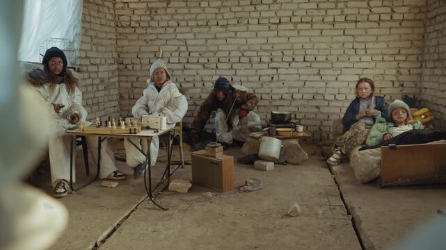 Stone age like survivors sit inside rundown shelter with cracked floor and tired faces, group tense as leader prepares to rise with cutlass, while makeshift kitchen and chessboard remain nearby
