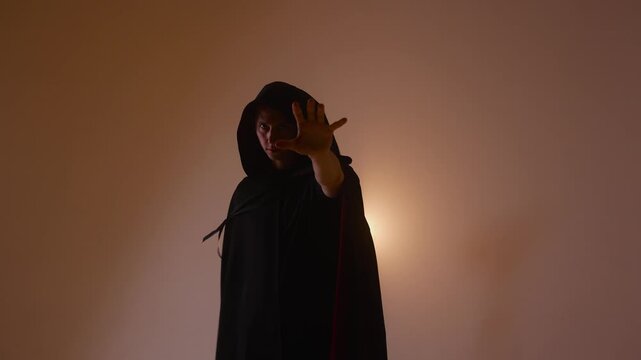 A cloaked and hooded stranger makes epic, sweeping gestures, backlit by a bright and warm light source, creating a powerful visual of a secretive mage or enigma.