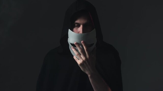 A young man in a mystical black hooded cloak puts a mask over his face, becoming a faceless stranger. Concept of concealing identity and secret society membership.