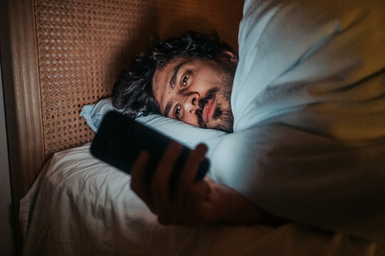 Man lying in bed at night, looking at his smartphone, suggesting themes of sleeplessness, technology use in the bedroom, or potential negative impact. 