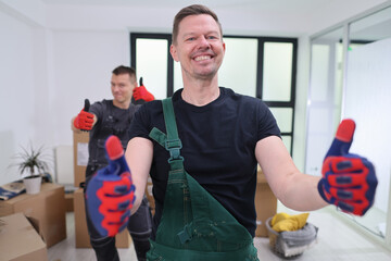 Two cheerful movers, one in green overalls and the other in a dark outfit, give thumbs up in a...