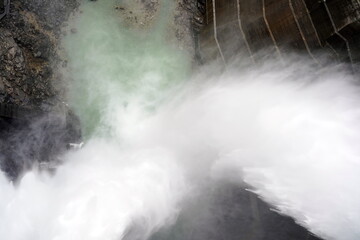 巨大なダムの放流口から大量の水が勢いよく流れ落ちる様子を真上から捉えた迫力ある風景。白い水しぶきが霧のように広がりエメラルドグリーンの川面へ落下する。