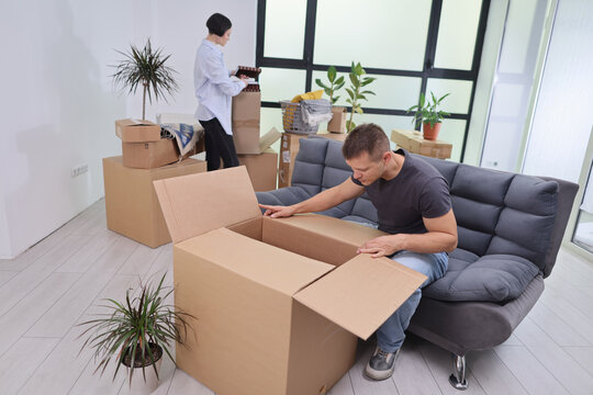 In a cozy, sunlit room, a couple takes a break from unpacking boxes. One person carefully explores a large box while the other arranges items among green plants and bright decor.