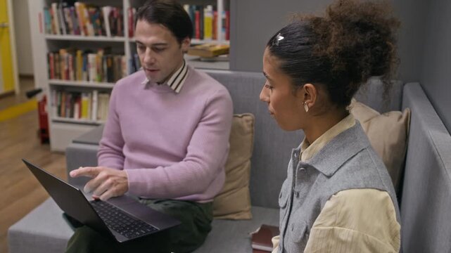 Zoom out shot of young diverse couple of motivated students coworking on their academic project or paper, sitting in comfortable modern library and studying together using digital technology and books
