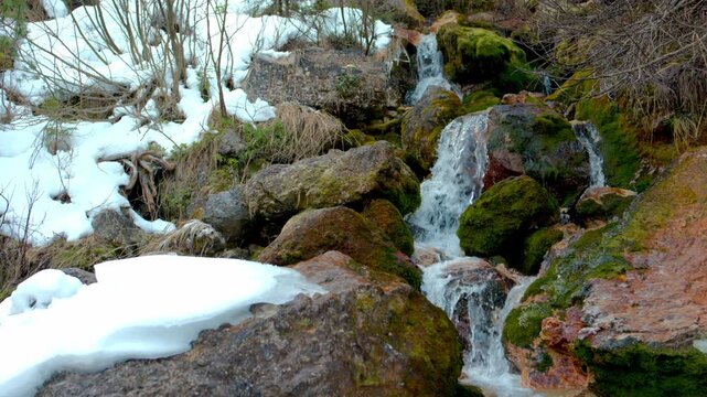 A clear spring flows between mossy rocks in slow motion, water falling gently while snow surrounds the stones in a cold, quiet mountain setting.