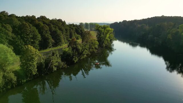 Aerial view of the Kolpa River near Metlika, Slovenia, with a lone canoeist paddling through calm water surrounded by lush green forest, reflected in the river during warm summer light.